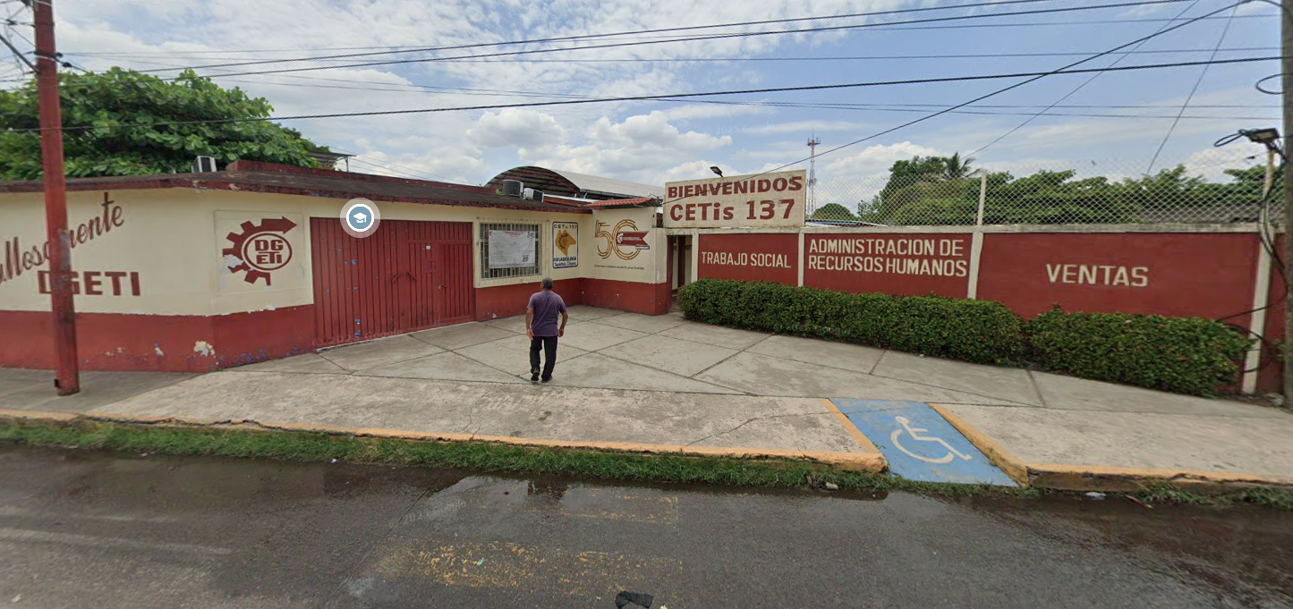Fotografía que muestra una explanada de forma triangular a la que se puede ingresar por medio de una rampa. La fachada del plantel está conformada por muros de color beige y rojo. Del lado derecho se muestra la puerta de acceso y encima hay un cartel que dice BIENVENIDOS CETIS 137.
